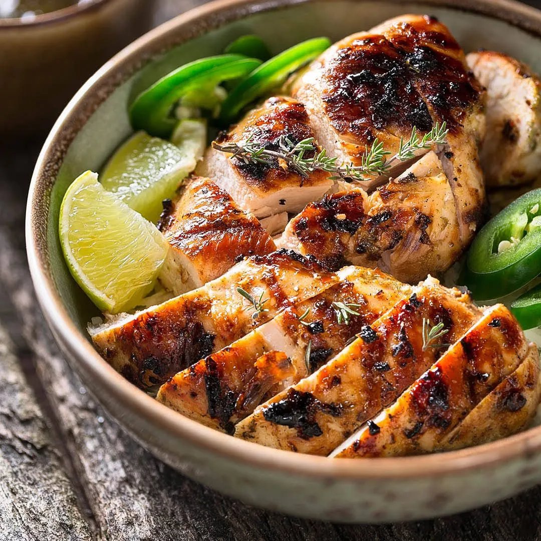 A bowl filled with sliced, grilled chicken breast, garnished with fresh herbs, lime wedges, and sliced jalapeƱos, placed on a rustic wooden surface.